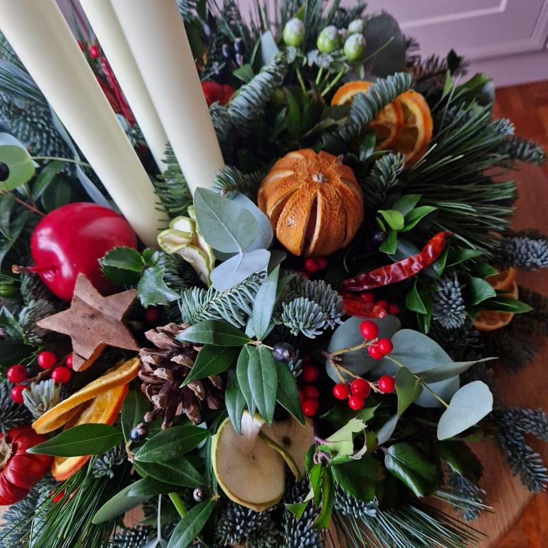 Traditional Christmas centrepiece with candles, fresh pine, holly, mixed foliage, dried fruits, chillies, oranges, apples, pine cones, pumpkins, and berries.