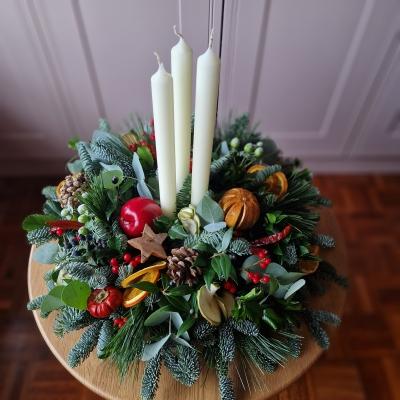 Traditional Christmas centrepiece with candles, fresh pine, holly, mixed foliage, dried fruits, chillies, oranges, apples, pine cones, pumpkins, and berries.