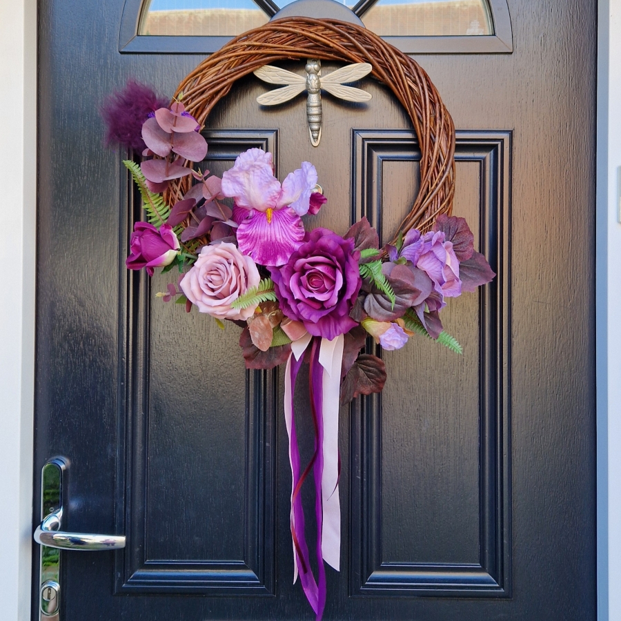 Handmade faux flower wreath in rich plum and pink tones, featuring flag iris, roses, and trailing ribbons, displayed on a black door with a metallic dragonfly ornament