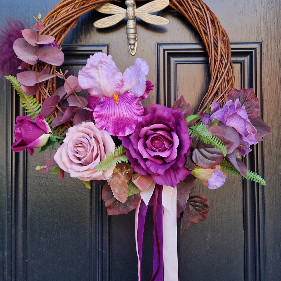 Handmade faux flower wreath in rich plum and pink tones, featuring flag iris, roses, and trailing ribbons, displayed on a black door with a metallic dragonfly ornament
