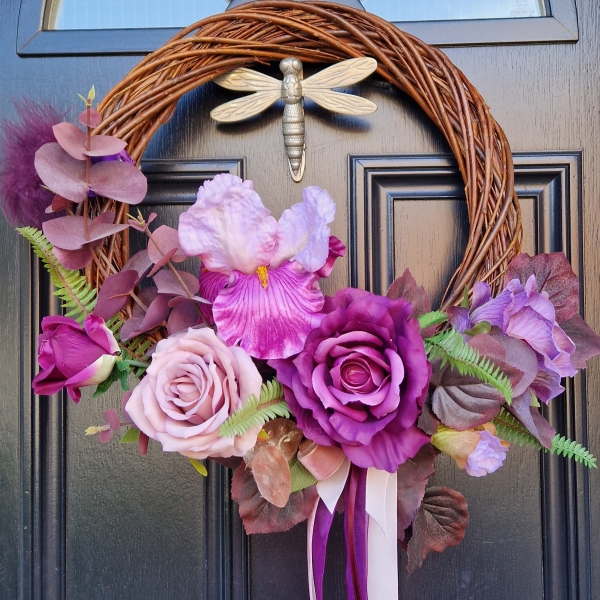 Handmade faux flower wreath in rich plum and pink tones, featuring flag iris, roses, and trailing ribbons, displayed on a black door with a metallic dragonfly ornament