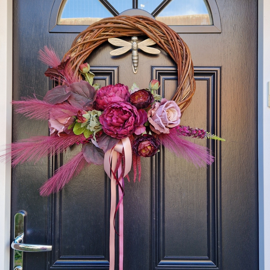 Decorative faux wreath on a black door, featuring deep damson and plum peonies, roses, layered ranunculus, and textured foliage arranged on a curved grapevine style base. Accented with trailing burgundy and pink ribbons, evoking a rich, romantic autumnal style.