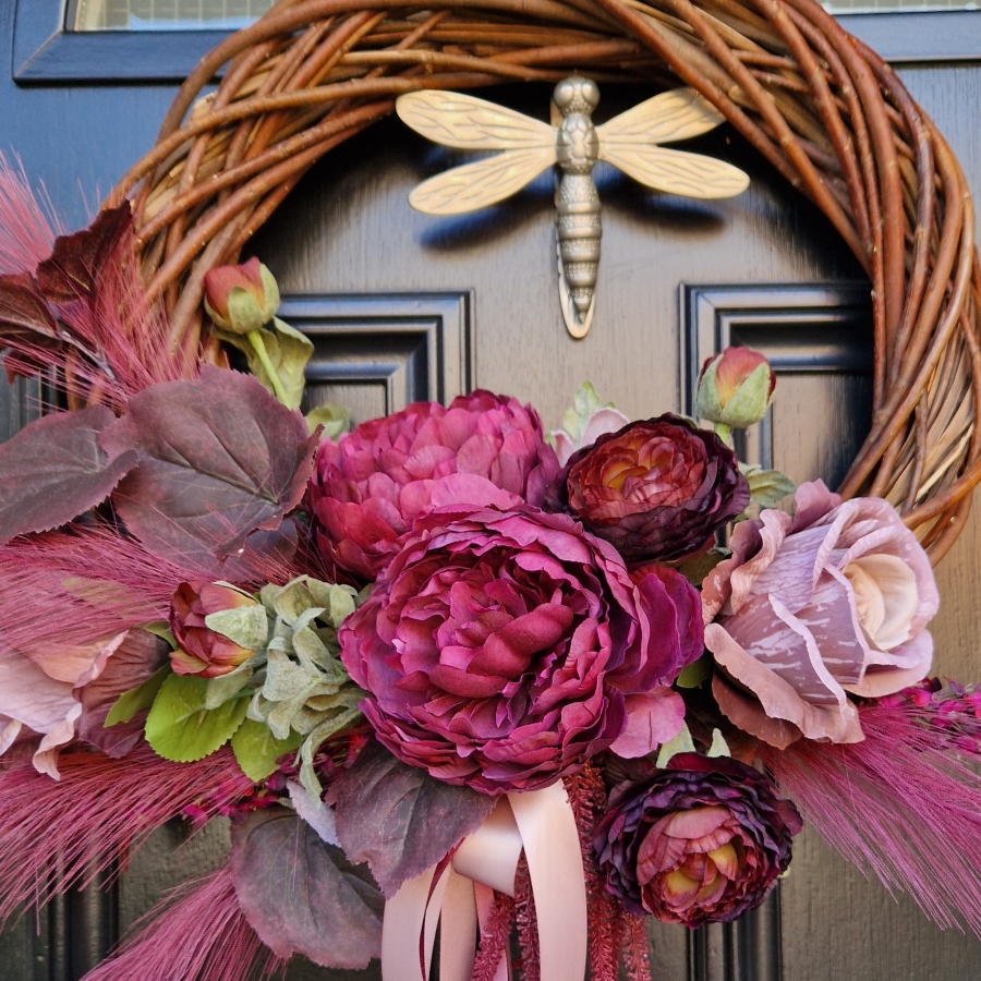 Decorative faux wreath on a black door, featuring deep damson and plum peonies, roses, layered ranunculus, and textured foliage arranged on a curved grapevine style base. Accented with trailing burgundy and pink ribbons, evoking a rich, romantic autumnal style.