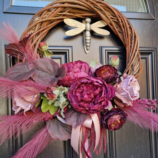 Decorative faux wreath on a black door, featuring deep damson and plum peonies, roses, layered ranunculus, and textured foliage arranged on a curved grapevine style base. Accented with trailing burgundy and pink ribbons, evoking a rich, romantic autumnal style.
