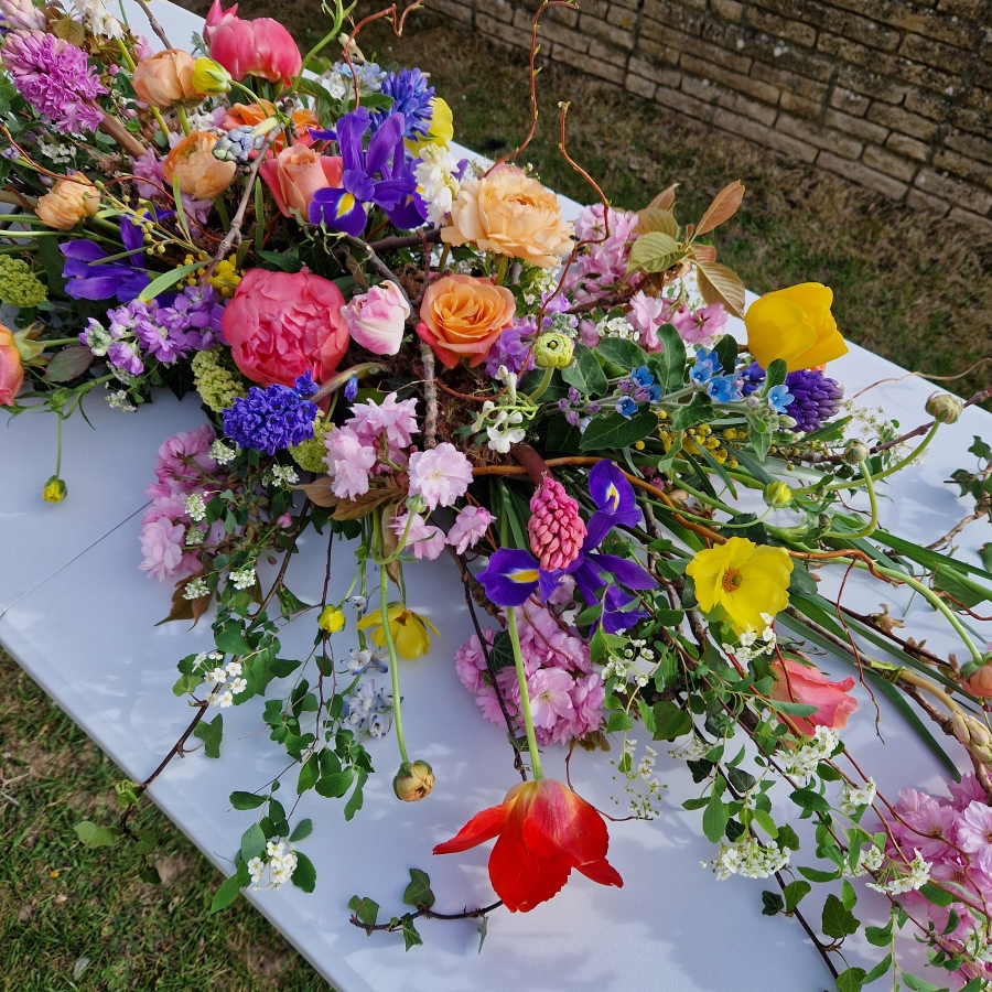 Foam‑free biodegradable coffin meadow with bright pink, apricot, yellow and purple seasonal flowers arranged on a visible moss, branch and chicken‑wire base.