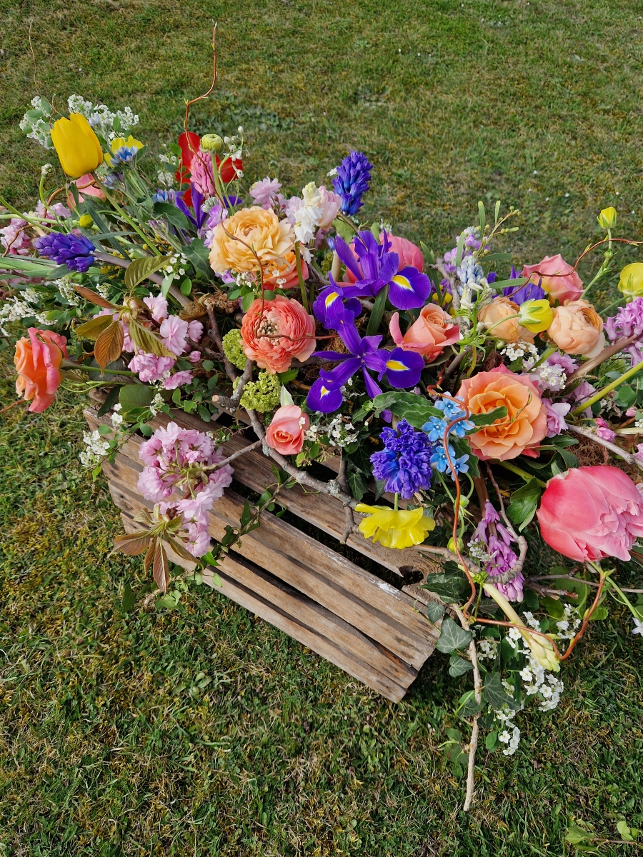 Foam‑free biodegradable coffin meadow with bright pink, apricot, yellow and purple seasonal flowers arranged on a visible moss, branch and chicken‑wire base.