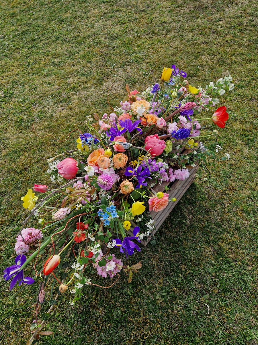 Foam‑free biodegradable coffin meadow with bright pink, apricot, yellow and purple seasonal flowers arranged on a visible moss, branch and chicken‑wire base.