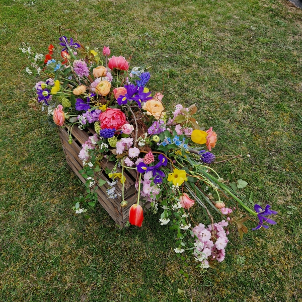 Foam‑free biodegradable coffin meadow with bright pink, apricot, yellow and purple seasonal flowers arranged on a visible moss, branch and chicken‑wire base.