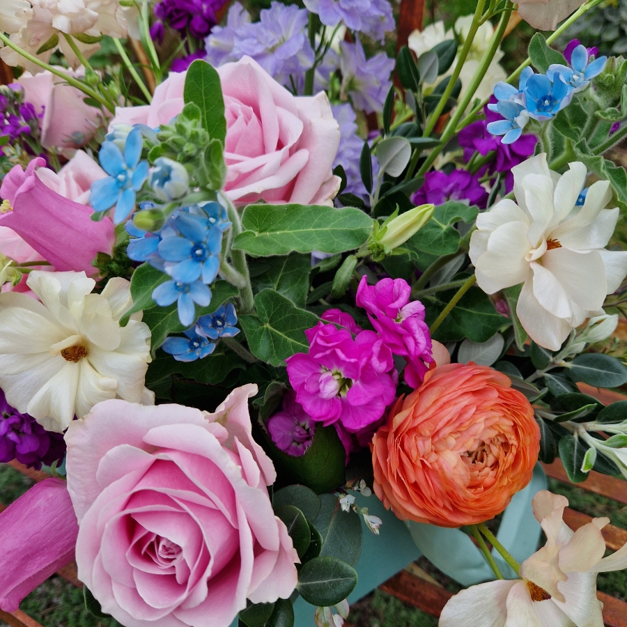 A pastel spring–summer hat box filled with premium blooms in soft pink, cream, apricot, blue and mauve, featuring roses, campanula, delphiniums, ranunculus and matthiola in a natural, garden style arrangement.