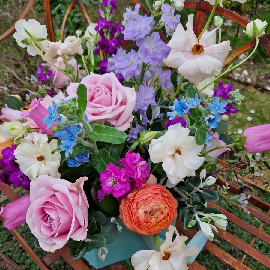 A pastel spring–summer hat box filled with premium blooms in soft pink, cream, apricot, blue and mauve, featuring roses, campanula, delphiniums, ranunculus and matthiola in a natural, garden style arrangement.