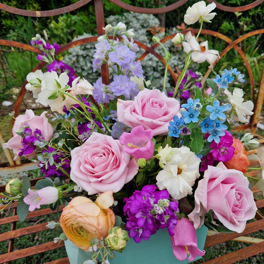 A pastel spring–summer hat box filled with premium blooms in soft pink, cream, apricot, blue and mauve, featuring roses, campanula, delphiniums, ranunculus and matthiola in a natural, garden style arrangement.