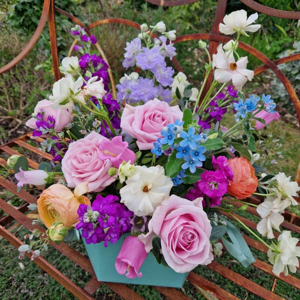 A pastel spring–summer hat box filled with premium blooms in soft pink, cream, apricot, blue and mauve, featuring roses, campanula, delphiniums, ranunculus and matthiola in a natural, garden style arrangement.