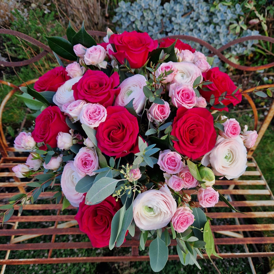 A hand‑tied Valentine’s bouquet of cerise and candy‑pink roses, pale pink ranunculus, camellia and eucalyptus, gift‑wrapped and presented in a kraft water‑holding bouquet box.