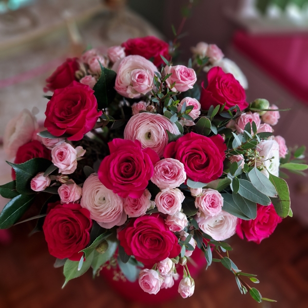 A hand‑tied Valentine’s bouquet of cerise and candy‑pink roses, pale pink ranunculus, camellia and eucalyptus, gift‑wrapped and presented in a kraft water‑holding bouquet box.