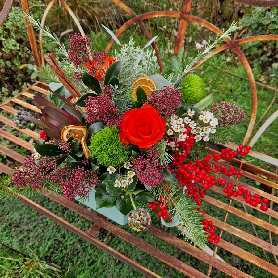 Christmas hat box arrangement with burnt orange roses, burgundy Skimmia, wax flower, ilex berries, pine, ivy berries, dried oranges, cinnamon sticks, and pine cones