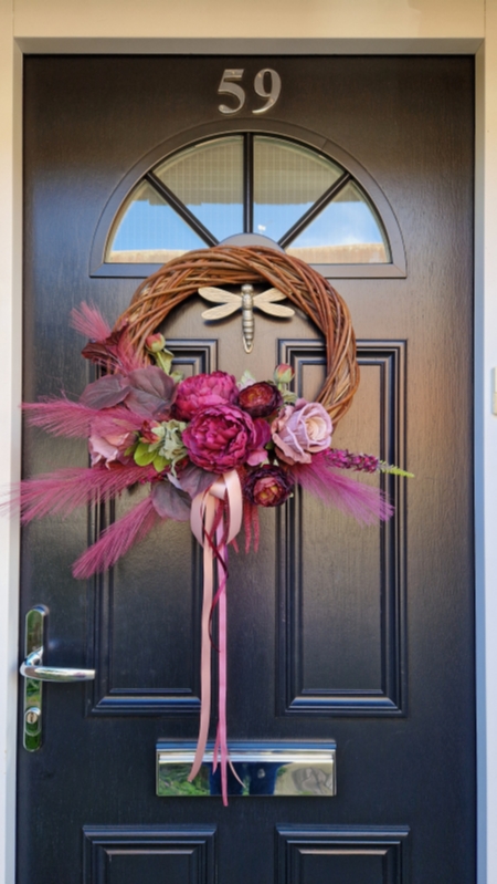 Decorative faux wreath on a black door, featuring deep damson and plum peonies, roses, layered ranunculus, and textured foliage arranged on a curved grapevine style base. Accented with trailing burgundy and pink ribbons, evoking a rich, romantic autumnal style.