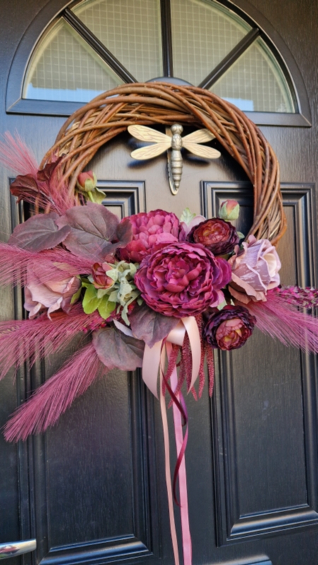 Decorative faux wreath on a black door, featuring deep damson and plum peonies, roses, layered ranunculus, and textured foliage arranged on a curved grapevine style base. Accented with trailing burgundy and pink ribbons, evoking a rich, romantic autumnal style.