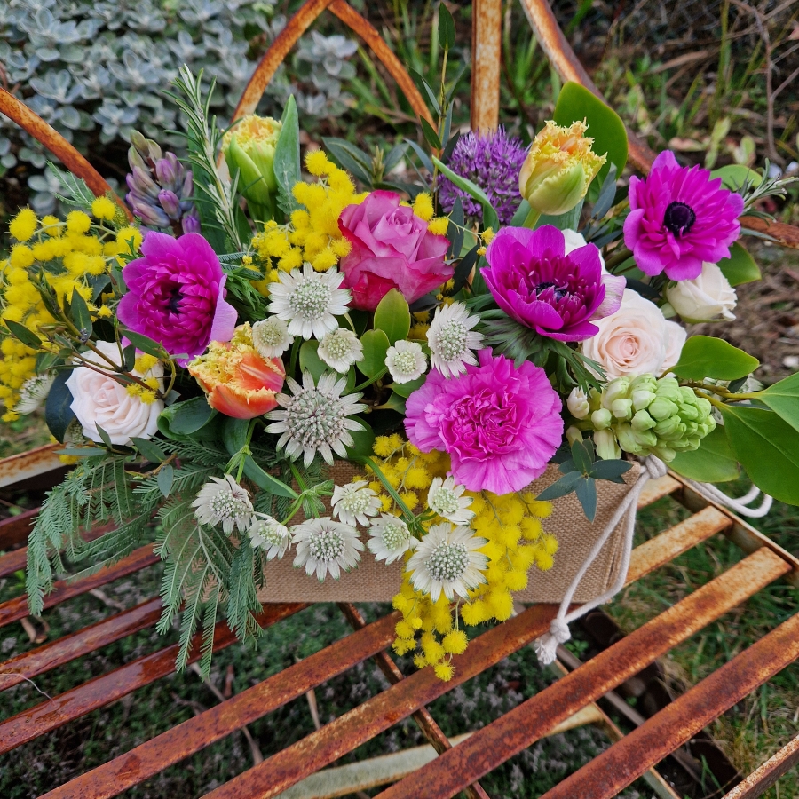 A spring arrangement in a jute trough-style box filled with mimosa, tulips, anemones, hyacinths, roses, allium, and carnations in apricot, fuchsia, yellow, and pink tones, arranged in a natural Floramay style.