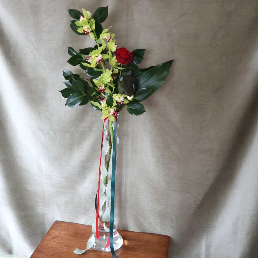 A tall glass vase arrangement featuring a green cymbidium orchid stem and a single velvety red rose, with glossy green foliage and trailing red and green ribbons.