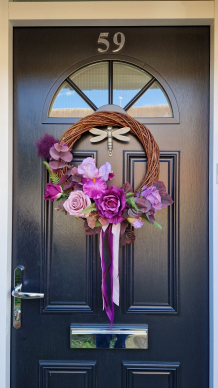 Handmade faux flower wreath in rich plum and pink tones, featuring flag iris, roses, and trailing ribbons, displayed on a black door with a metallic dragonfly ornament