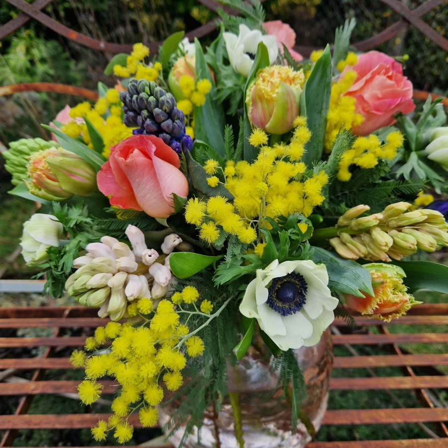 A spring bouquet in a blush glass vase featuring mimosa, tulips, anemones, roses, and hyacinths in apricot, yellow, pink, and fuchsia tones, arranged in a loose, natural Floramay style.