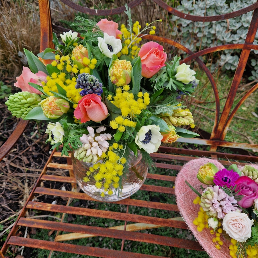 A spring bouquet in a blush glass vase featuring mimosa, tulips, anemones, roses, and hyacinths in apricot, yellow, pink, and fuchsia tones, arranged in a loose, natural Floramay style.