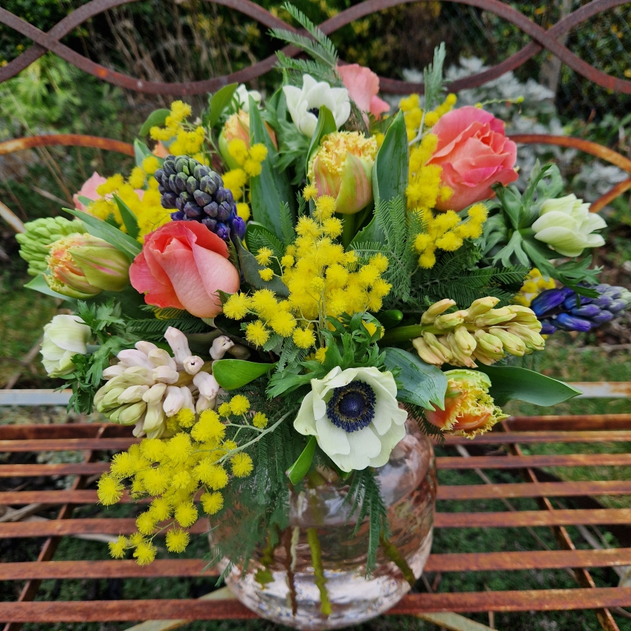 A spring bouquet in a blush glass vase featuring mimosa, tulips, anemones, roses, and hyacinths in apricot, yellow, pink, and fuchsia tones, arranged in a loose, natural Floramay style.
