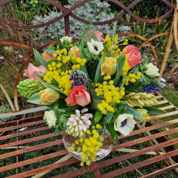 A spring bouquet in a blush glass vase featuring mimosa, tulips, anemones, roses, and hyacinths in apricot, yellow, pink, and fuchsia tones, arranged in a loose, natural Floramay style.