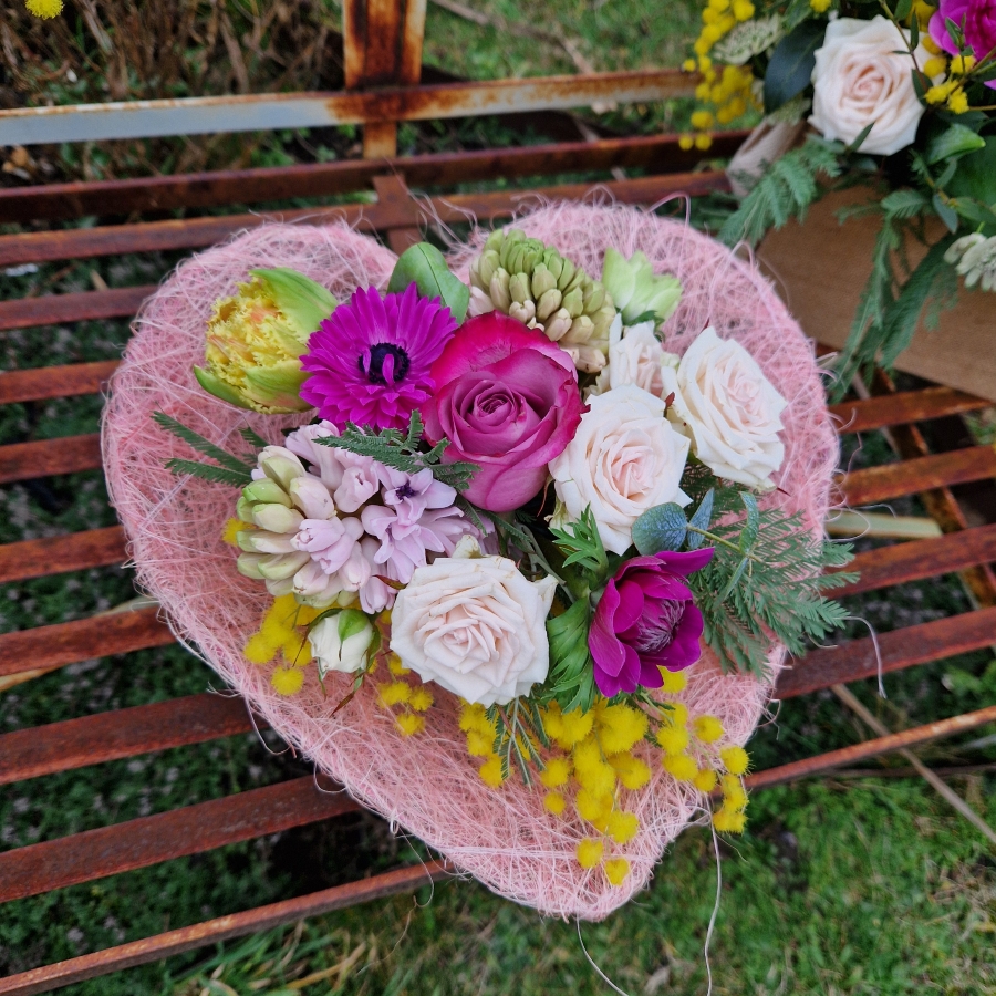 A small spring bouquet in a pink glass jam jar, edged with a pink sisal heart and filled with mimosa, tulips, anemones, roses, and hyacinths in apricot, yellow, pink, and fuchsia tones, arranged in a natural Floramay style.