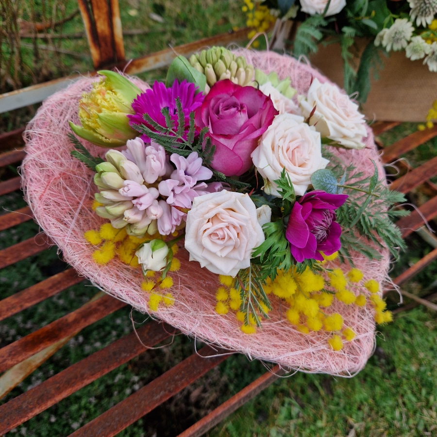 A small spring bouquet in a pink glass jam jar, edged with a pink sisal heart and filled with mimosa, tulips, anemones, roses, and hyacinths in apricot, yellow, pink, and fuchsia tones, arranged in a natural Floramay style.