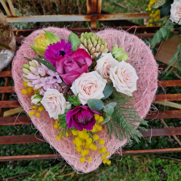 A small spring bouquet in a pink glass jam jar, edged with a pink sisal heart and filled with mimosa, tulips, anemones, roses, and hyacinths in apricot, yellow, pink, and fuchsia tones, arranged in a natural Floramay style.