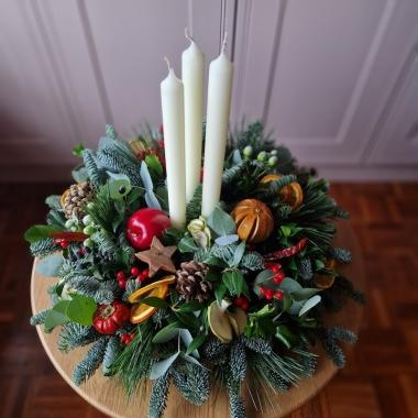 Traditional Christmas centrepiece with candles, fresh pine, holly, mixed foliage, dried fruits, chillies, oranges, apples, pine cones, pumpkins, and berries.
