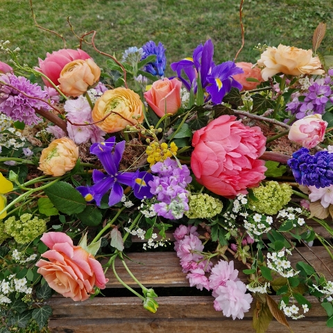 Foam‑free biodegradable coffin meadow with bright pink, apricot, yellow and purple seasonal flowers arranged on a visible moss, branch and chicken‑wire base.