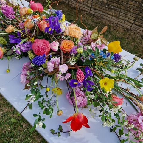 Foam‑free biodegradable coffin meadow with bright pink, apricot, yellow and purple seasonal flowers arranged on a visible moss, branch and chicken‑wire base.