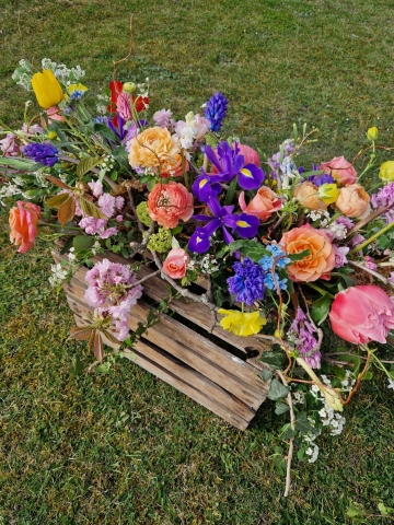 Foam‑free biodegradable coffin meadow with bright pink, apricot, yellow and purple seasonal flowers arranged on a visible moss, branch and chicken‑wire base.