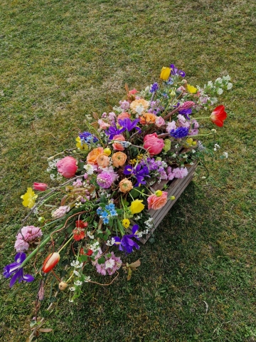 Foam‑free biodegradable coffin meadow with bright pink, apricot, yellow and purple seasonal flowers arranged on a visible moss, branch and chicken‑wire base.