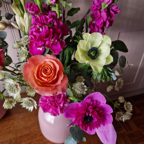 A spring arrangement in a matt pink ceramic vase featuring matthiola, tuberose, allium, roses, carnations, and anemones in fuchsia, apricot, yellow, and pink tones, styled in a natural Floramay design.