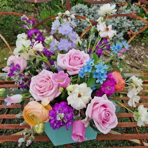 A pastel spring–summer hat box filled with premium blooms in soft pink, cream, apricot, blue and mauve, featuring roses, campanula, delphiniums, ranunculus and matthiola in a natural, garden style arrangement.