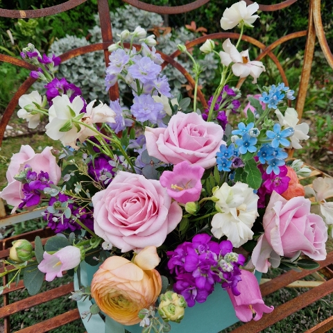 A pastel spring–summer hat box filled with premium blooms in soft pink, cream, apricot, blue and mauve, featuring roses, campanula, delphiniums, ranunculus and matthiola in a natural, garden style arrangement.