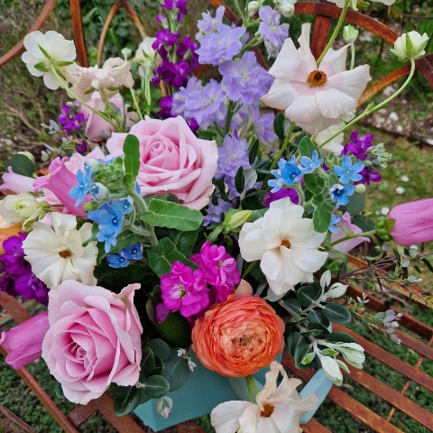 A pastel spring–summer hat box filled with premium blooms in soft pink, cream, apricot, blue and mauve, featuring roses, campanula, delphiniums, ranunculus and matthiola in a natural, garden style arrangement.