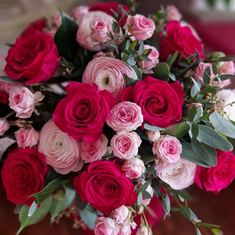 A hand‑tied Valentine’s bouquet of cerise and candy‑pink roses, pale pink ranunculus, camellia and eucalyptus, gift‑wrapped and presented in a kraft water‑holding bouquet box.