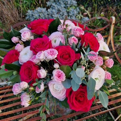 A hand‑tied Valentine’s bouquet of cerise and candy‑pink roses, pale pink ranunculus, camellia and eucalyptus, gift‑wrapped and presented in a kraft water‑holding bouquet box.