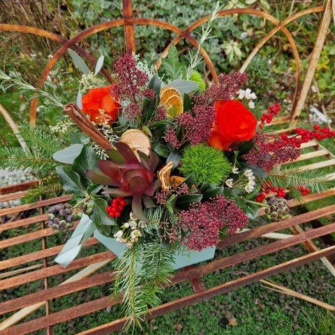 Christmas hat box arrangement with burnt orange roses, burgundy Skimmia, wax flower, ilex berries, pine, ivy berries, dried oranges, cinnamon sticks, and pine cones