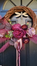 Decorative faux wreath on a black door, featuring deep damson and plum peonies, roses, layered ranunculus, and textured foliage arranged on a curved grapevine style base. Accented with trailing burgundy and pink ribbons, evoking a rich, romantic autumnal style.