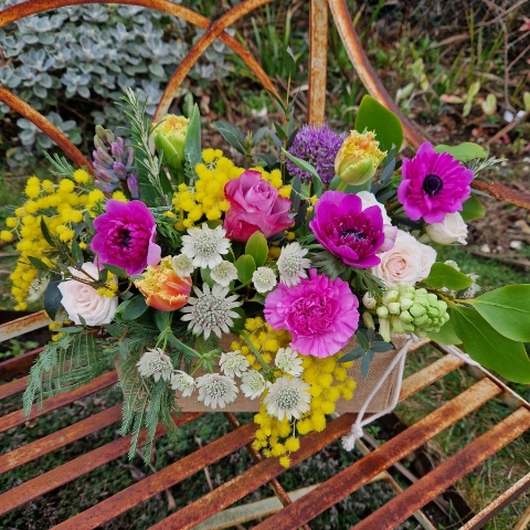 A spring arrangement in a jute trough-style box filled with mimosa, tulips, anemones, hyacinths, roses, allium, and carnations in apricot, fuchsia, yellow, and pink tones, arranged in a natural Floramay style.