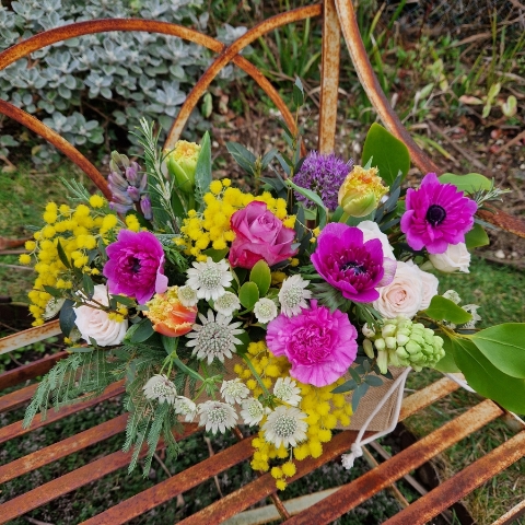 A spring arrangement in a jute trough-style box filled with mimosa, tulips, anemones, hyacinths, roses, allium, and carnations in apricot, fuchsia, yellow, and pink tones, arranged in a natural Floramay style.