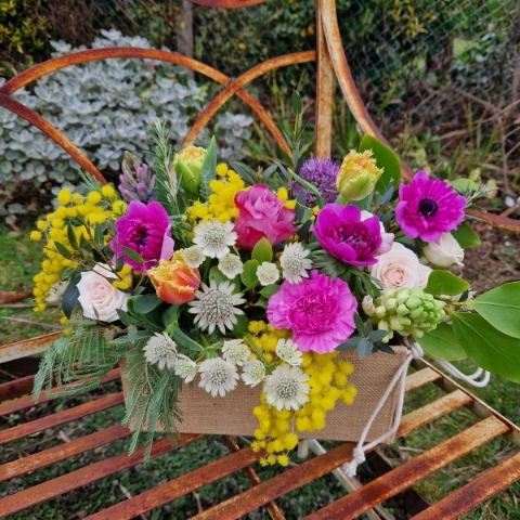 A spring arrangement in a jute trough-style box filled with mimosa, tulips, anemones, hyacinths, roses, allium, and carnations in apricot, fuchsia, yellow, and pink tones, arranged in a natural Floramay style.