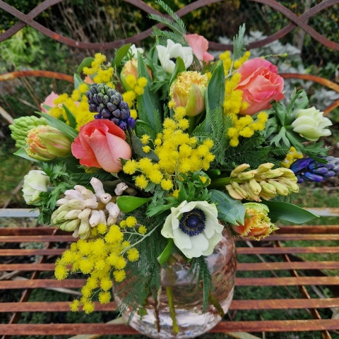 A spring bouquet in a blush glass vase featuring mimosa, tulips, anemones, roses, and hyacinths in apricot, yellow, pink, and fuchsia tones, arranged in a loose, natural Floramay style.