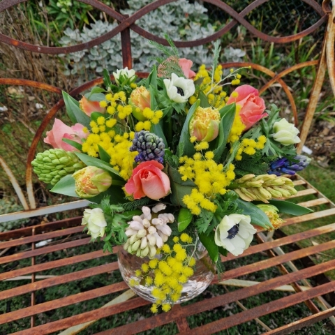 A spring bouquet in a blush glass vase featuring mimosa, tulips, anemones, roses, and hyacinths in apricot, yellow, pink, and fuchsia tones, arranged in a loose, natural Floramay style.