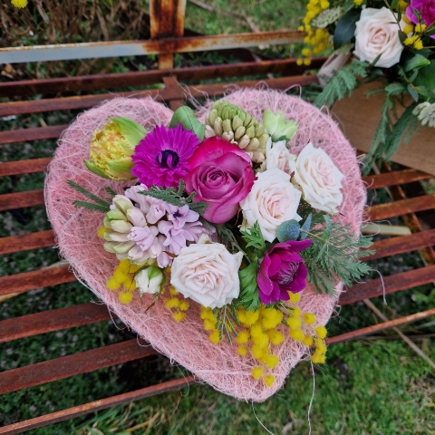 A small spring bouquet in a pink glass jam jar, edged with a pink sisal heart and filled with mimosa, tulips, anemones, roses, and hyacinths in apricot, yellow, pink, and fuchsia tones, arranged in a natural Floramay style.
